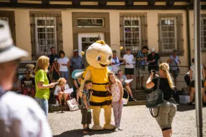 Maskottchen in Bienenkostüm beim Landes-Musik-Festival posiert mit zwei Kindern – fröhliche Festivalatmosphäre mit Publikum im Hintergrund.